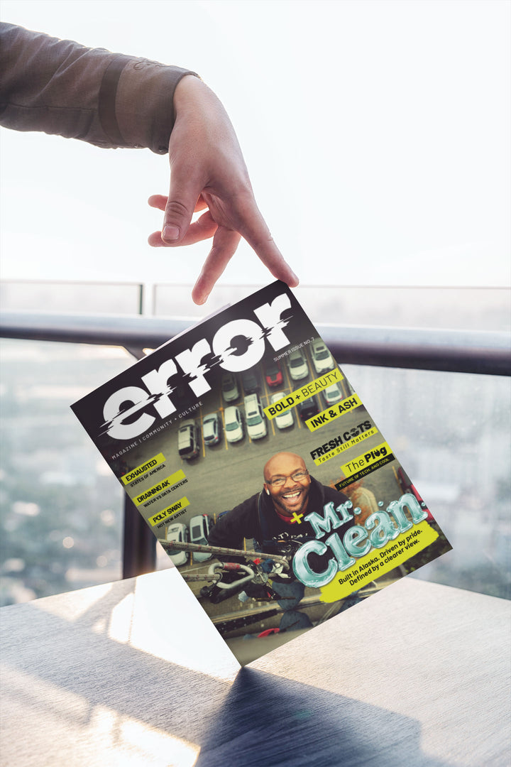 A hand points at a tilted copy of Error magazine on a table. The magazine cover features a smiling man and the headline 'Mr.
