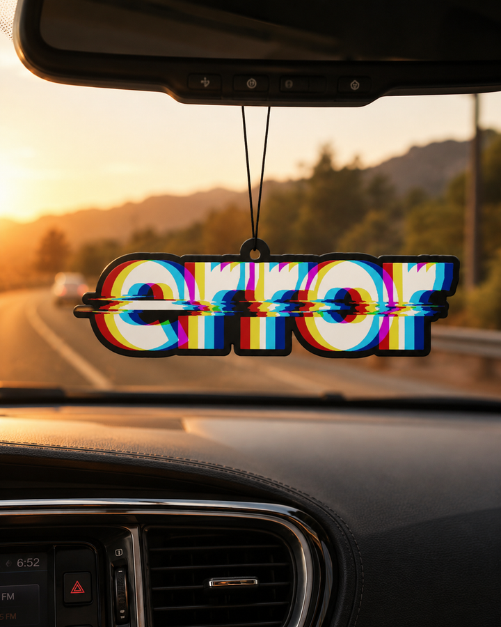 A holographic 'ERROR' air freshener hanging from a rearview mirror inside a car, with a blurred sunset road scene in the background.