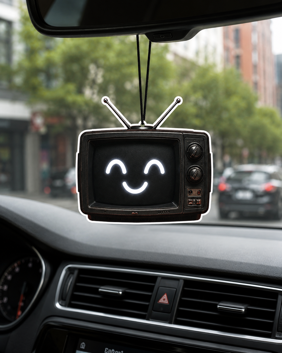 A retro-style television car air freshener or ornament hanging from a rearview mirror inside a car. The TV has a black screen with a white glowing smiley face and two silver antennas, set against a blurred background of a city street.