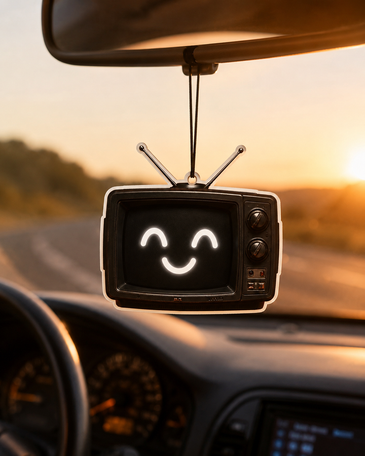 A retro-style hanging television air freshener with a smiling face on the screen, suspended from a car's rearview mirror on a road trip at sunset.