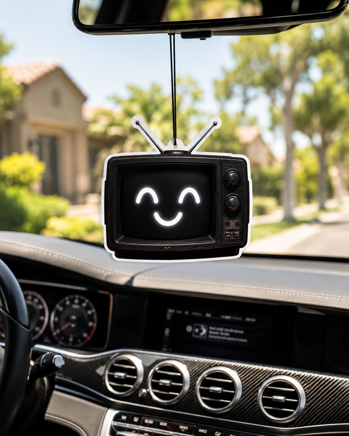 A retro-style television car air freshener hanging from a rearview mirror in a car interior, with a smiling face displayed on its screen and a blurred suburban street background visible through the windshield.